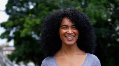 Caddede kameraya poz verirken gülümseyen ve gülen mutlu bir Brezilyalı genç kadın. Kıvırcık saçlı, Afro-Amerikan kökenli, otantik, neşeli.