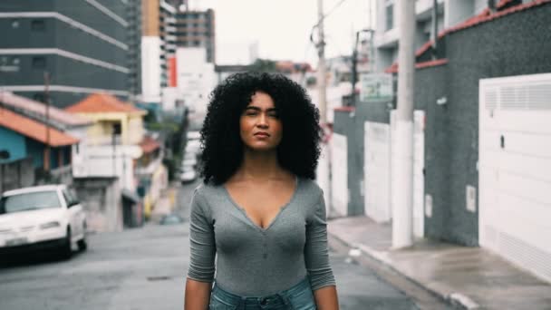都市の外に立っているカーリーな髪を持つ自信のある若い黒人女性は 厳粛な表情でカメラを見ています 20代 ストリートポートレート顔 — ストック動画