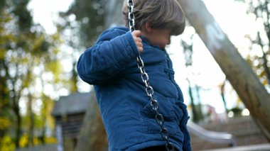 Sonbahar gününde mavi ceketle salıncağa tırmanmaya çalışan bir çocuk. 3 yaşındaki küçük bir çocuk salıncağın tepesinde oturmaya çalışıyor. Deneme yanılma konsepti.