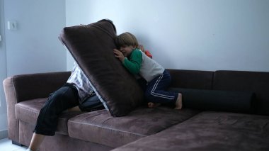 Çocuk kanepede koşuyor annesinin yastık kalesine dalıyor, gerçek bir aile zamanı, anne ve oğlu evde birlikte oynuyorlar.