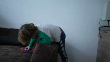 Küçük bir çocuk evde oynarken kanepenin kenarına tırmanıyor. Çocuk kanepede oturuyor.