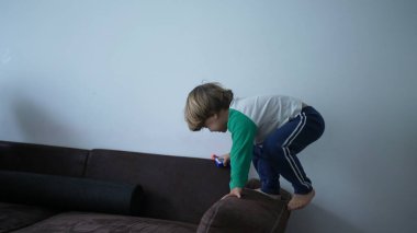 Küçük bir çocuk evde oynarken kanepenin kenarına tırmanıyor. Çocuk kanepede oturuyor.