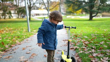 Küçük bir çocuk, sonbahar günü parkta dikilip mavi ceketini giyiyor ve 3 tekerlekli scooterını kontrol ediyor.