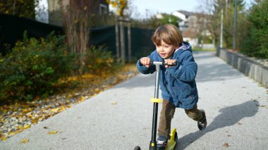 Neşeli 3 yaşındaki çocuk Sonbahar Günışığı 'nda dışarıda oyuncak scooter sürüyor.
