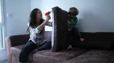 Çocuk kanepede koşuyor annesinin yastık kalesine dalıyor, gerçek bir aile zamanı, anne ve oğlu evde birlikte oynuyorlar.