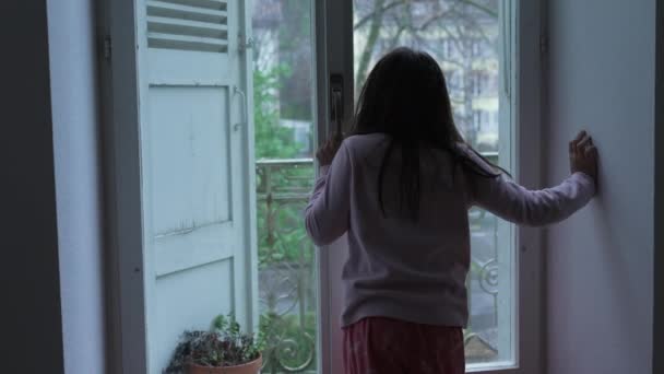 沉思的孩子的后背 站在窗边 从二楼的公寓里 忧郁而忧郁地凝视着风景 有思想的小女孩靠在玻璃上 — 图库视频影像