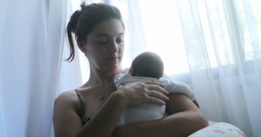 Mom holding newborn baby next to window curtain