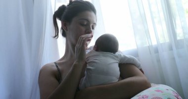 Mom holding newborn baby next to window curtain