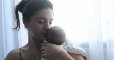 Mother holding newborn baby next to home window, first week of life