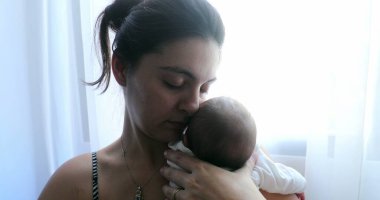 Mother holding newborn baby next to home window, first week of life