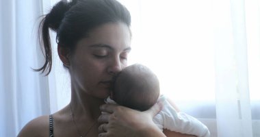 Mother holding newborn baby next to home window, first week of life