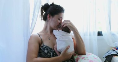 Mother holding newborn baby in her arms next to home window