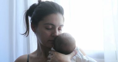 Mother holding newborn baby next to home window, first week of life