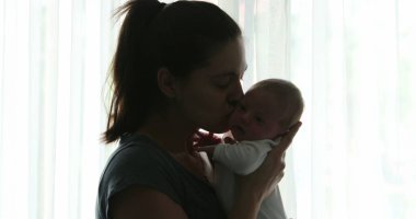 Mother kissing newborn baby infant next to window curtain