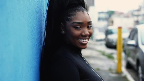 一位快乐的非洲裔美国年轻女子 在城市街道上靠着蓝色的墙壁 微笑着转过头对着摄像机追踪拍摄到的运动镜头 — 图库视频影像
