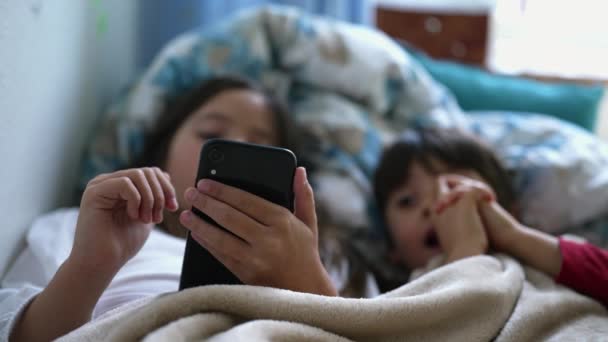 特写小女孩的手在床底下的床单下拿着手机装置 小弟弟在旁边看着屏幕 两个从事现代技术的孩子 — 图库视频影像