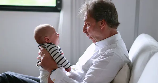 Candid Grand-father holding newborn grand-son baby in sofa indoors authentic grandpa interaction