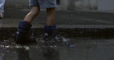 Çocuk su birikintisinde yürüyor kaldırımda yağmur botları giyiyor süper yavaş çekimde 800 fps her yere damlacıklar saçıyor