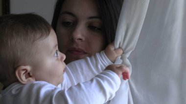Anne kucağında bebek, perde penceresiyle oynuyor.
