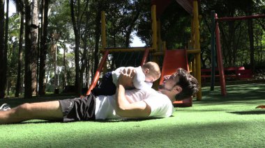 Baba, çocuk parkında bebeği havaya kaldırıyor.