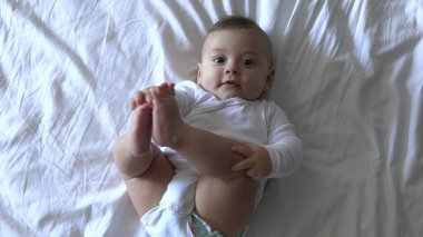 Yatakta tatlı bir erkek bebek kameraya bakıp gülümsüyor ve ayaklarını tutuyor.