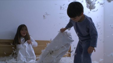 Çocuklar akşamları yastık kavgasının ortasında tüylerle oynuyorlar. Küçük çocuk, pijama giymiş kız kardeşine tüy atıyor.