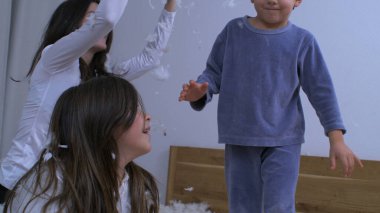 Çocuklar yastık tüyleriyle 1000 fps hızla süper yavaş çekimde oynuyorlar. Anne ve kız, otantik mutlu anda küçük kardeşine kuş tüyü fırlatıyorlar.