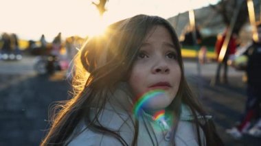 Gün batımında, arka planda parkın önünde parlayan, arka planda parlayan, bulanık yüzlü 8 yaşındaki çocuklar oynayan küçük bir kız çocuğu.