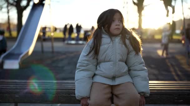 冬の夕暮れの間 公園のベンチに座った熟考の少女は バックグラウンドプレーでぼやけた子供たちとコートを着ていました フレアでバックライト 静かなペンギン8歳の子供 思考で失われた — ストック動画