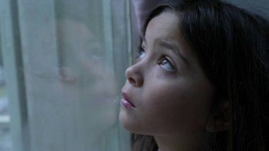 Üzgün, düşünceli çocuk camdan dışarı bakıyor ve kendini evde kapana kısılmış hissediyor. Yalnızlık çeken küçük bir kız