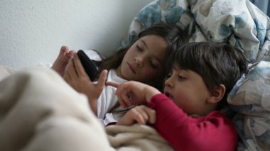 Çocuklar yatak çarşaflarının altında yatarken cep telefonu cihazına bakıyorlar. Kardeşler modern eğlence medyası tarafından özümsenip telefon ekranına bakıyorlar.