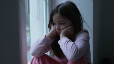 Üzgün, düşünceli, küçük bir kız. Pencerenin kenarında yalnızlık hissedip düşünceli bakışlarla manzaraya bakıyor. Derin düşüncelere dalmış çocuk