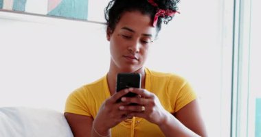 Evdeki koltukta oturan cep telefonu cihazına bakan genç siyah Brezilyalı bir kadın. 20 'li yaşlarda Afrika kökenli bir kadın elinde telefonla ekrana bakıyor ve uyarı mesajı okuyor.