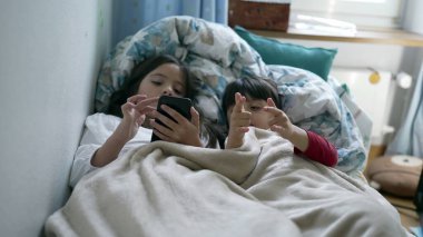 Çocuklar yatak örtüsünün altında cep telefonu ekranını paylaşıyorlar. Küçük kız ve erkek kardeş modern teknolojiyi kullanıyor.