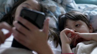 Çocuklar yatakta uzanmış telefon ekranına bakıyor, erkek ve kız kardeşlerinin yakın çekimleri çevrimiçi içeriği izliyor, eğlence medyası tarafından özümseniyor.