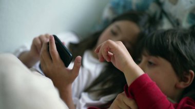 Genç Kardeşler Yatakta Cep Telefonu Ekranını Paylaşıyorlar, Kız Kardeş ve Erkek Kardeş Akıllı Telefona Bakıyorlar
