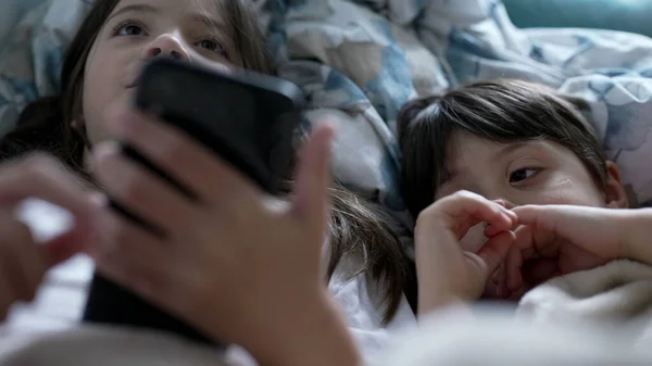 Children looking at phone screen - sister holding cellphone while small brother stares at media online, kids lying in bed under bedsheets engaged with modern technology