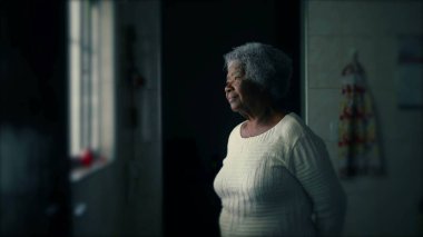 Düşünceli, yaşlı, 80 'li yaşlarda bir kadın mütevazı bir evde, pencerenin önünde düşünceli bir ifadeyle dikiliyor. Gri saçlı, yalnız, düşünceli yaşlı bayan.