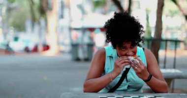 Genç siyahi bir kadın parkta hamburger yiyor. 20 'li yaşlarda Afrika kökenli bir kadın güneşli bir günde öğle yemeğinde fast-food yiyor.