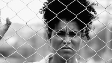 Metal parmaklıklara yaslanmış kızgın bir siyah kadın, hayatın zorluklarıyla boğuşurken yüzünü buruşturarak kameraya bakıyor.