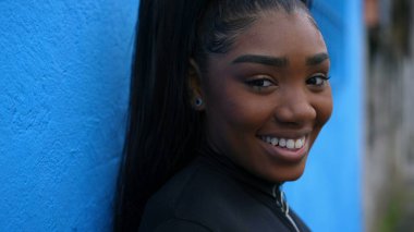 Güney Amerika 'da şehir ortamında mavi duvara yaslanmış kameraya gülümseyen 20' li yaşlardaki mutlu siyah Latin bir kızın portresi. Afrika kökenli genç bir kadın izleme çekiminde kameraya doğru dönüyor.