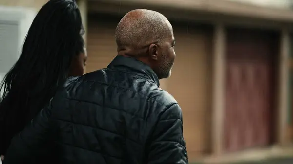 Tracking shot of a middle-aged father walking with his teenage daughter outside in urban street during drizzle rain. African American mature person bonding with family while walking forward