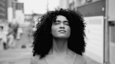 Umutlu, genç, kıvırcık saçlı bir kadın sokakta dikilip gökyüzüne bakıyor. Siyah beyaz UMUT ve FAITH ile birlikte. Afrikalı Amerikalı Tanrı 'nın varlığını hissediyor.