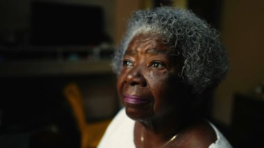 Kıdemli Afro-Amerikan bir kadının kapalı alanda yalnız başına pencereye baktığı yakın çekim yüzü. Düşünceli, yaşlı, beyaz saçlı, düşünceli bir zenci kadın.