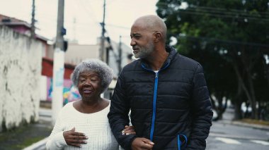 Orta yaşlı Afrikalı Amerikalı bir adam 80 yaşındaki annesiyle şehir sokaklarında gezintiye çıkıyor.