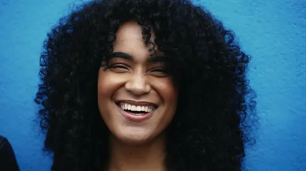 One joyful young latina black woman with curly hair smiling at camera on blue backdrop in city environment. close-up face of South American person of African descent