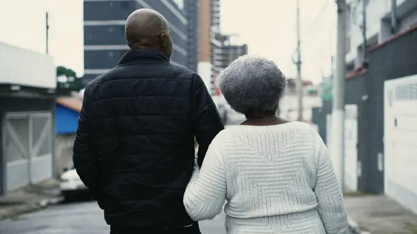 Back of African American Adult Son Walking with Elderly gray hair Mother in City, Senior Lady in Her 80s Arm-in-Arm with Caretaker Outdoors during drizzle
