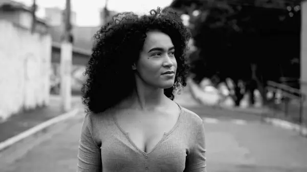One confident young black woman walking towards camera outside in urban street environment in intense monochromatic, black and white. Curly hair African American 20s girl