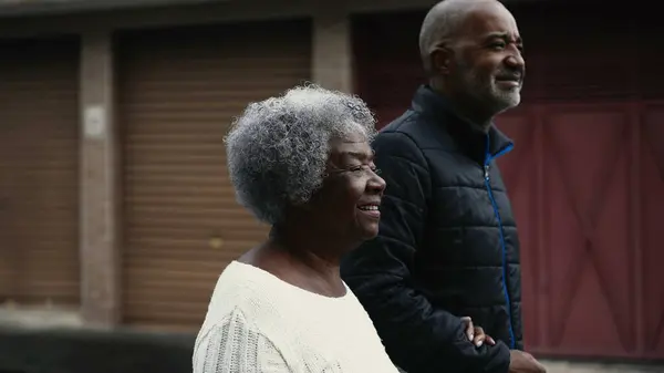 Joyful South American black Elderly gray hair lady in 80s walking in street with the help of her adult middle aged son with arm around during daily stroll in urban environment