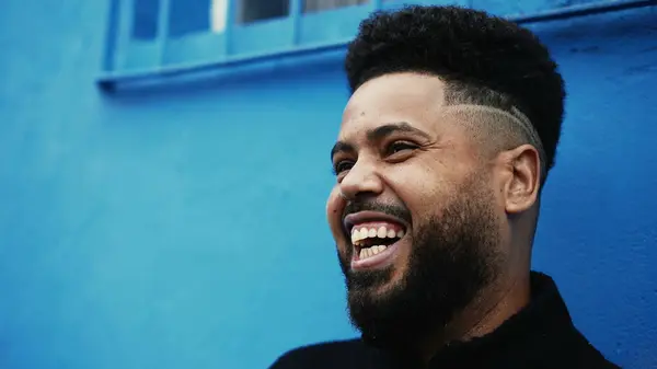 One happy young black Brazilian man laughing and smiling in front of an urban blue wall, close-up face of a South American person conversing with friends off-camera